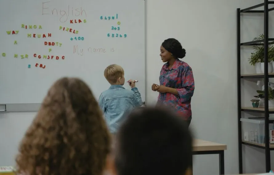 Criança escreve no quadro enquanto professora observa em sala de aula.