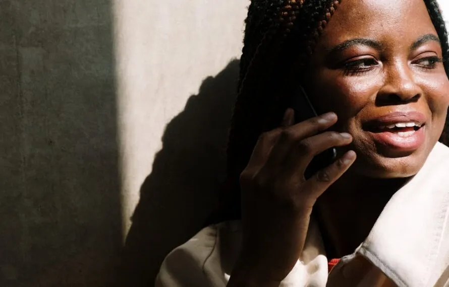 Automação do WhatsApp: Do Chat à Conversão e Como Vender Mais com Estratégias Inteligentes Mulher com cabelos trançados sorrindo enquanto fala ao telefone com a mão ao rosto