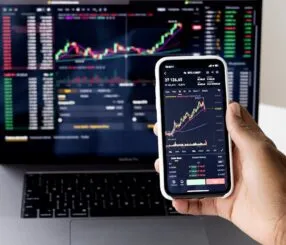 Pessoa observando gráficos de criptomoedas no celular e no laptop.