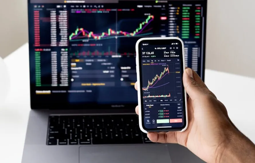 Pessoa observando gráficos de criptomoedas no celular e no laptop.