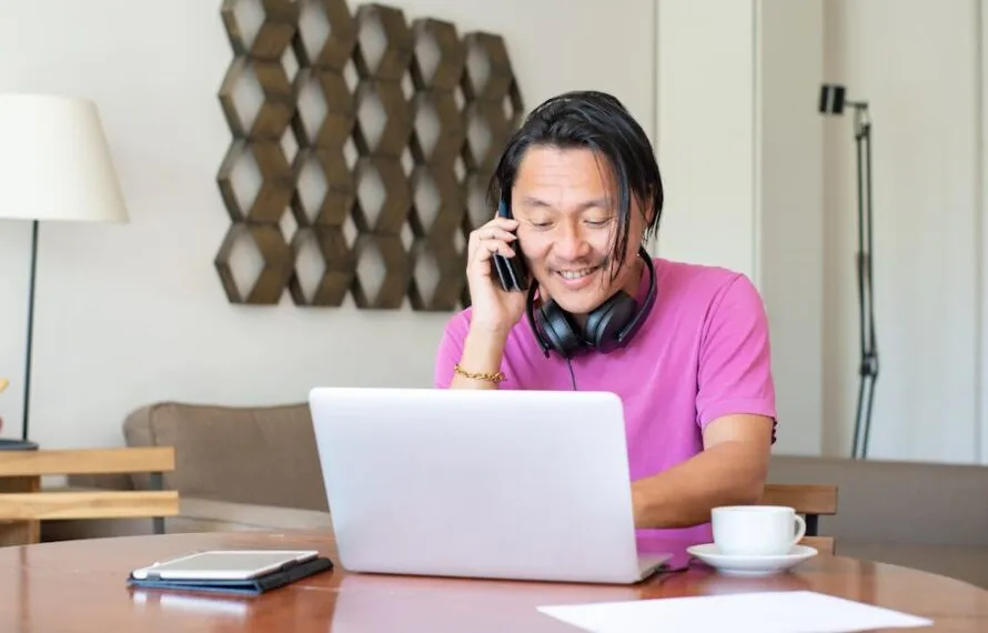 Homem com fones de ouvido falando ao telefone enquanto usa um laptop em casa.