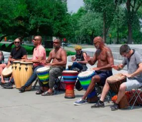 Como a musicalização ajuda adultos a aprender música do zero Grupo de adultos de diferentes idades tocando djembês e tambores numa roda de percussão ao ar livre em um parque